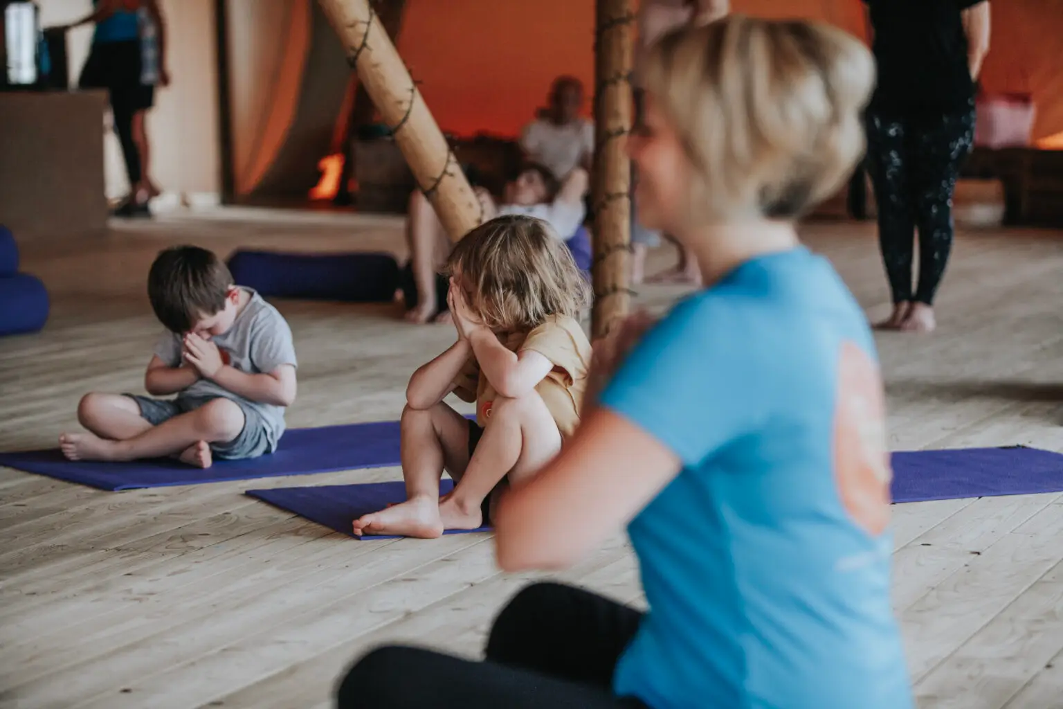 Children sitting cross-legged on yoga mats with their hands together as a Happy Kids Yoga teacher leads the class
