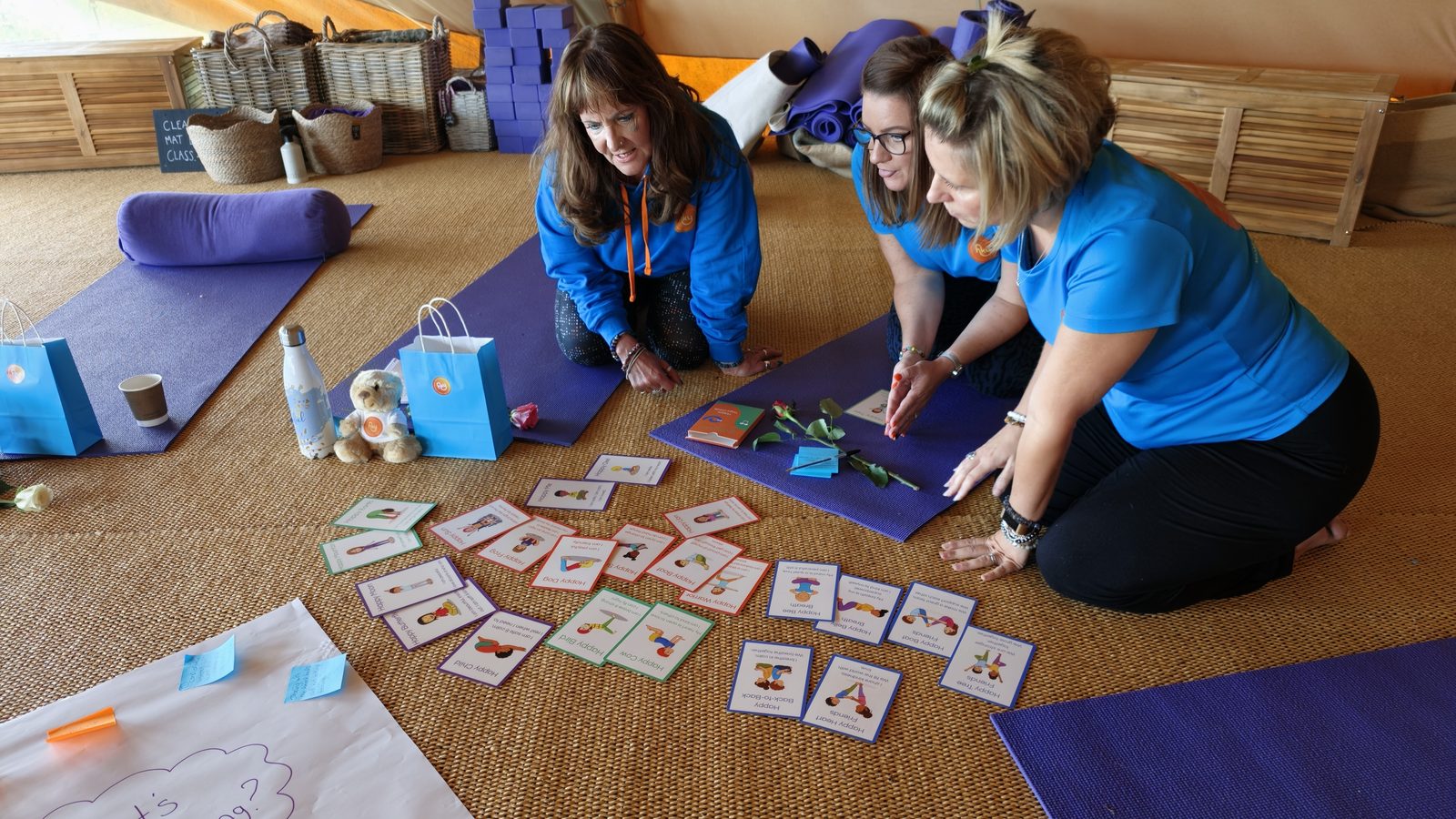 Happy Kids Yoga franchisees looking at the new Happy Kids Yoga pose cards laid out on a mat, with blue goodie bags beside them